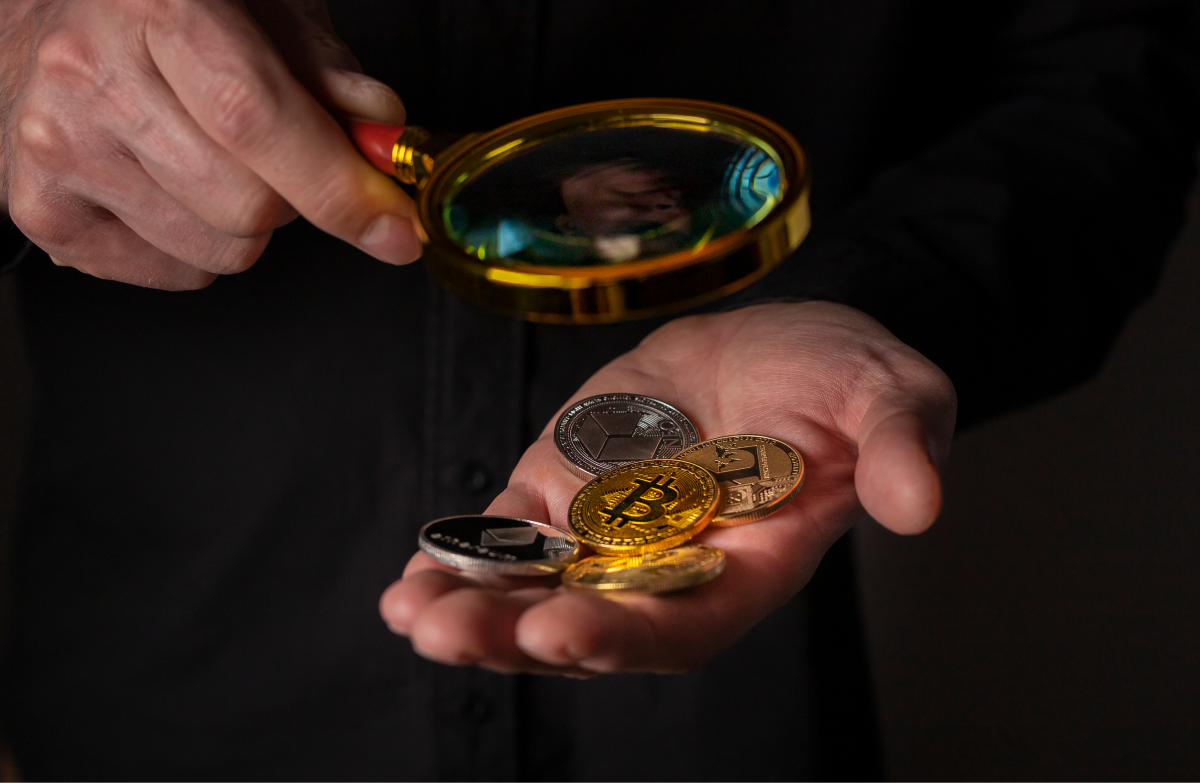 A hand holds a handful of cryptocurrency while another holds a magnifying glass over it