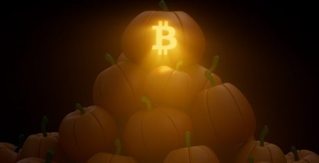 Sitting atop a pile of pumpkins is one carved with the Bitcoin logo. The top pumpkin is lit like a jack-o'-lantern.