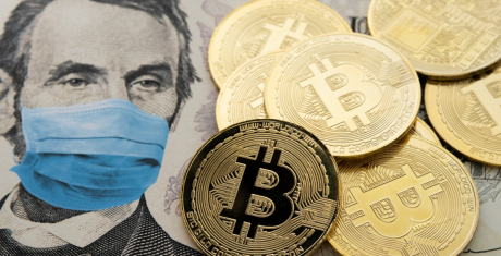 Several gold coins with the Bitcoin logo sit on a five-dollar bill. The portrait of President Abraham Lincoln on the bill is wearing a surgical mask. The relationship between cryptocurrency and covid is implied.